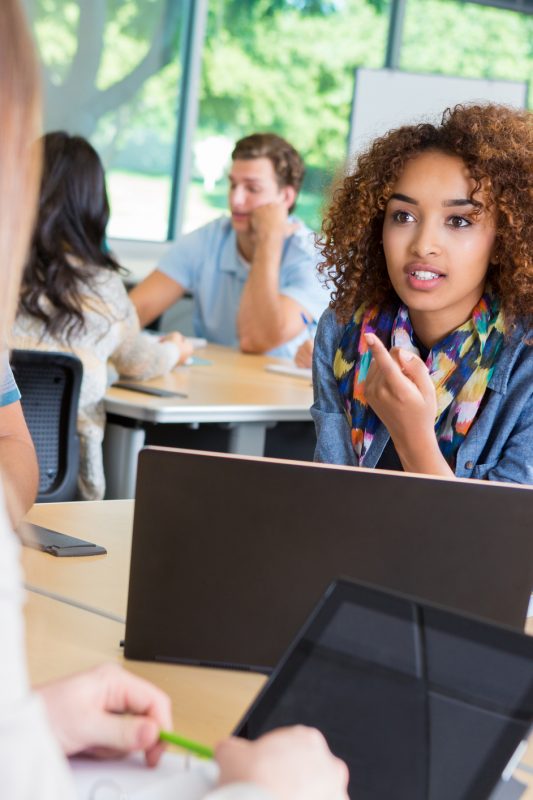 Young adult or teenage African American woman is student in college. She is discussing assignment and brainstorming during study session with diverse classmates in modern classroom. They are dressed in trendy casual clothing, and are using laptops and digital tablets.